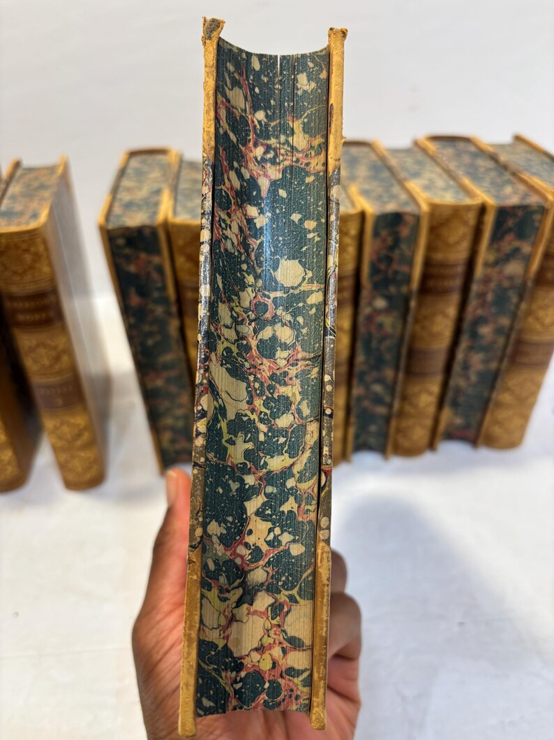 May include: A collection of antique books with aged, textured covers. The spines are a marbled pattern of teal, cream, and red. The edges are gilded, and the covers are a warm, aged tan color. The books are likely from a vintage collection.