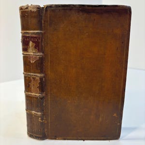May include: An antique leather-bound book with a brown cover and a raised spine. The spine features a dark red label with the text M'LAURIN'S SERMONS in gold lettering. The leather shows signs of wear, including scuffs and surface abrasions.