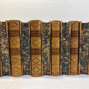May include: A collection of antique books with brown leather covers and gold lettering. The spines feature the title "Emerson's Works" and decorative gold patterns. The edges of the pages have a marbled design in shades of green, pink, and brown.