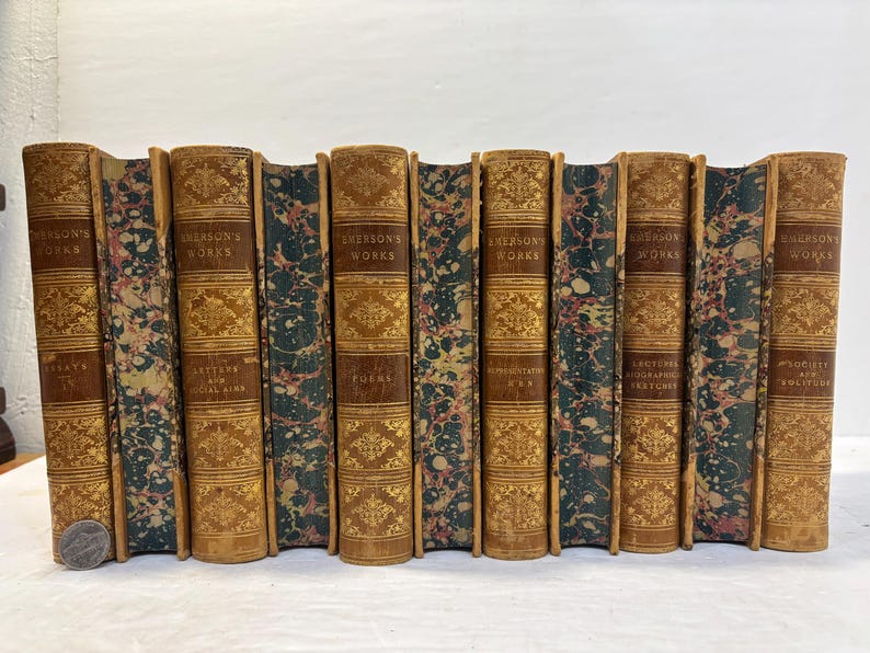 May include: A collection of antique books with brown leather covers and gold lettering. The spines feature the title "Emerson's Works" and decorative gold patterns. The edges of the pages have a marbled design in shades of green, pink, and brown.
