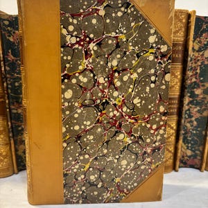 May include: A collection of antique books with leather-bound spines and marbled covers. The featured book has a tan leather spine and corner accents, with a marbled pattern in shades of gray, white, red, and yellow. The edges of the pages are visible.