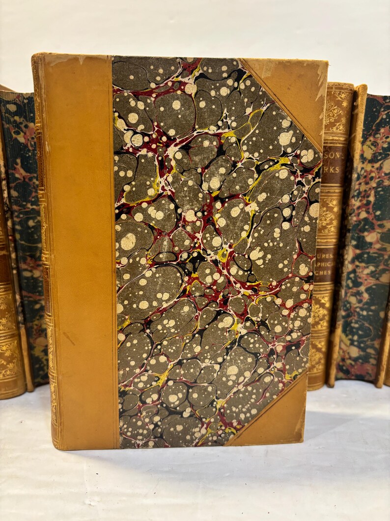 May include: A collection of antique books with leather-bound spines and marbled covers. The featured book has a tan leather spine and corner accents, with a marbled pattern in shades of gray, white, red, and yellow. The edges of the pages are visible.
