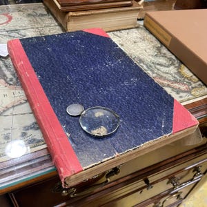 May include: A vintage, dark blue, hardback book with red edges. The book is lying on a wooden surface with a magnifying glass and a silver coin on top of it.