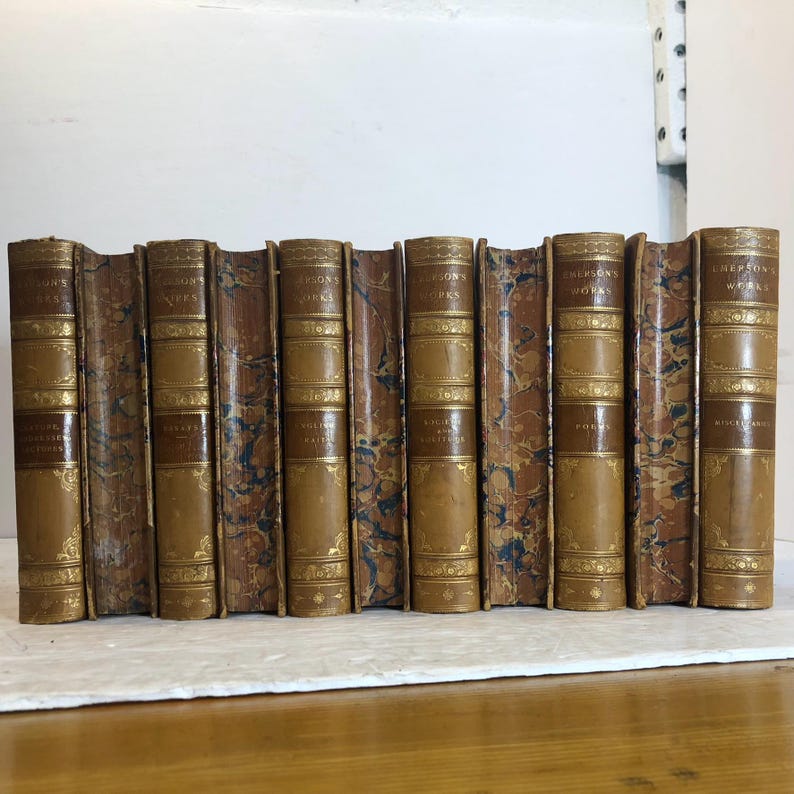 May include: A collection of antique books with brown leather spines and covers. The books are embossed with gold lettering and decorative patterns. The titles "Emerson's Works" are visible on the spines. The books are arranged in a row, suggesting a library or collection.