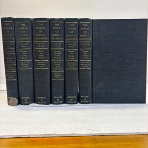 May include: A collection of six dark blue hardcover books titled "The Works of John C. Calhoun." The spines display volume numbers and titles, with gold lettering. The books are arranged side by side, with a seventh book of the same color to the right.