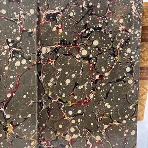 May include: Open antique book with marbled endpapers. The endpapers feature a dark gray background with a swirling pattern of black, red, yellow, and white. The book's spine is visible on the right.