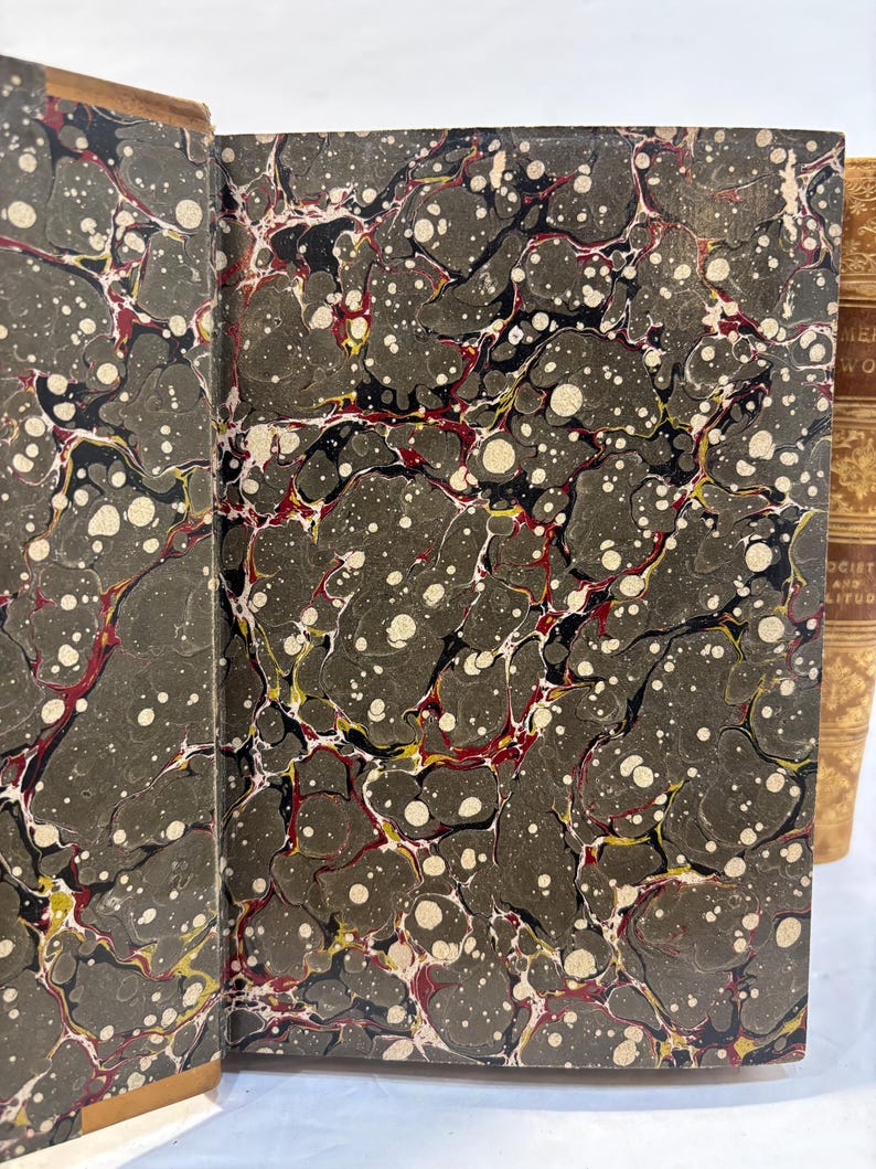 May include: Open antique book with marbled endpapers. The endpapers feature a dark gray background with a swirling pattern of black, red, yellow, and white. The book's spine is visible on the right.