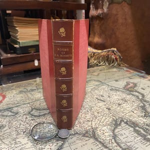May include: A red leather-bound book titled "Poems of D.G. Rossetti" with gold lettering and decorative rose illustrations on the spine. The book is sitting on a map.