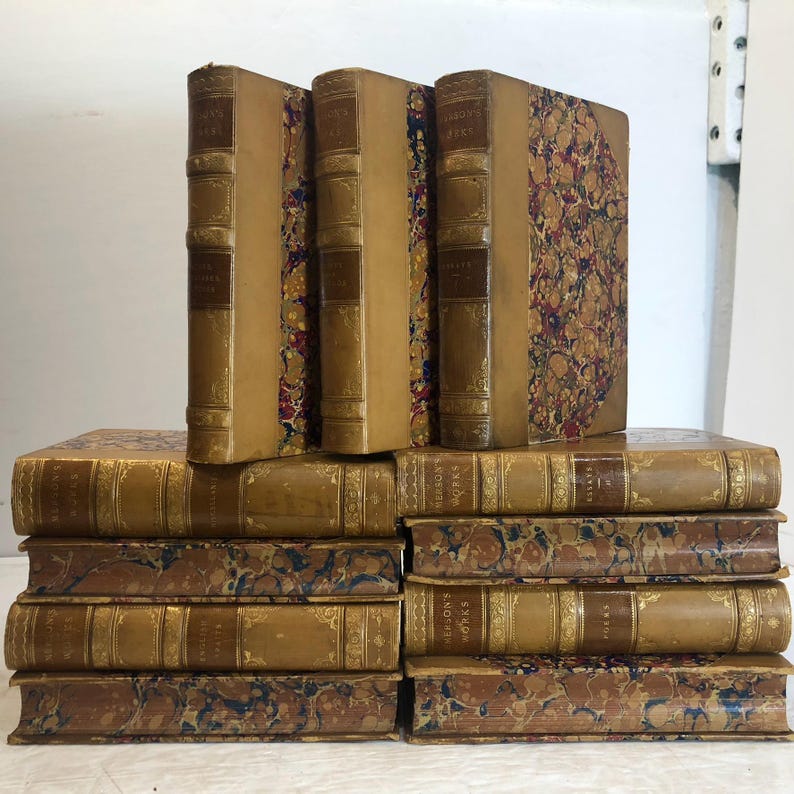 May include: A collection of antique books with tan leather spines and covers. The books have gold lettering and decorative marbled endpapers. Some books are stacked, while others stand upright.