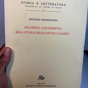 May include: A vintage book titled "STORIA E LETTERATURA" with the author Arnaldo Momigliano. The cover is cream-colored with black and red text. The book is part of the "RACCOLTA DI STUDI E TESTI" series, published in Rome in 1984.