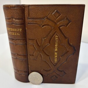 May include: A brown leather-bound book with gold lettering on the spine reading METHODIST HYMNS and on the front cover reading BECKIE MORGART. The cover features an embossed geometric pattern. A silver US quarter is placed at the base for scale.