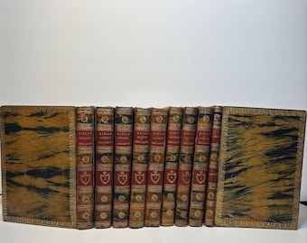 May include: A collection of antique books with leather-bound covers. The spines are red and gold with ornate detailing. The covers are a marbled brown and black pattern. The books are arranged in a row, with the covers open on either side.