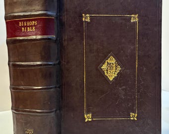 May include: A dark brown leather-bound book titled Bishops Bible on the spine with gold lettering. The spine features raised bands and the year 1573 in gold. The front cover has a gold-tooled rectangular border and a central diamond-shaped emblem.