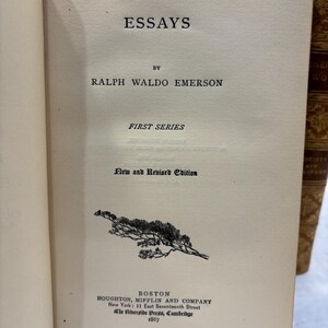 May include: Open book with the title "ESSAYS" by Ralph Waldo Emerson. The cover is off-white with black text and a small illustration. The book is a "New and Revised Edition" published in Boston in 1887.