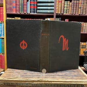May include: A black hardcover book with the title "Mussolini" in gold lettering on the spine. The front cover features a red "M" in a stylized font. The back cover has a red symbol with a sword and wreath.