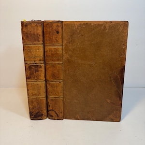 May include: Three antique books with worn, brown leather covers. The spines of the two books on the left have gold lettering. The right book is closed, showing a plain, textured cover. A yellow bookmark is visible at the top.