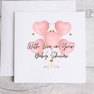 May include: A white baby shower card with a matching envelope. The card features five pink heart-shaped balloons with gold ribbons and star accents. The text "With Love on Your Baby Shower" is written in black script.