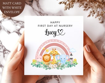 Może przedstawiać: Składana matowa kartka o wymiarach 15 cm x 15 cm z białą kopertą, zawierająca napis Happy First Day At Nursery oraz imię Lucy. Projekt zawiera akwarelową ilustrację różowej tęczy, lwa, żyrafy, słonia oraz zebry.