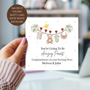May include: A 15.24x15.24 cm folded matte card with a white envelope featuring a line of baby items including overalls, a teddy bear, a onesie, and a bib hanging from red heart clips. Text reads: You're Going To Be Amazing Parents, Congratulations on your Exciting News, Melissa & John.