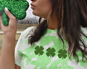May include: A woman wearing a green and white tie-dye shirt with four green shamrock designs. She is holding a large green glitter shamrock.