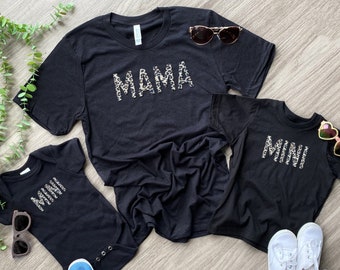 May include: A set of three black t-shirts with the words "Mama" and "Mini" in leopard print. The shirts are laid out on a wooden surface.