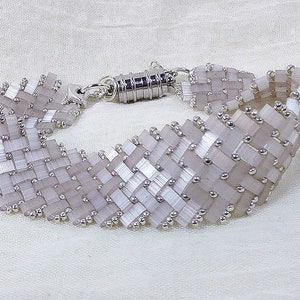 Puede incluir: Un brazalete blanco y plateado hecho de pequeñas cuentas cuadradas con un cierre plateado.