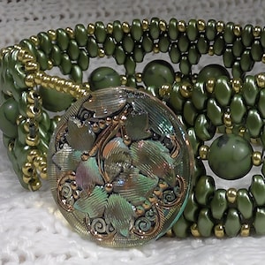 Può includere: Un braccialetto largo con perline verde oliva con un bottone decorativo rotondo in stile antico. Il braccialetto presenta perline verde oliva con dettagli dorati e perline verde oliva più grandi. Il bottone ha un design floreale con toni verdi e bronzo.