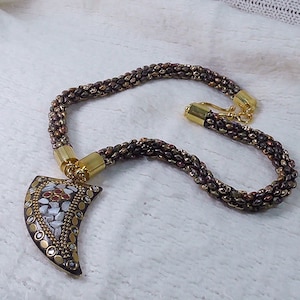 Può includere: Una collana di perline marroni con una chiusura dorata e un grande ciondolo dorato ornato a forma di dente.
