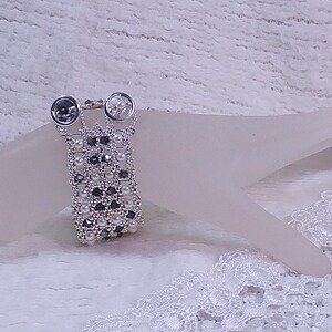 Può includere: Un anello argentato con due pietre rotonde e trasparenti e un elemento centrale rettangolare con perline. L'elemento centrale è decorato con piccole perline chiare e grigio scuro. L'anello è presentato su una forma astratta rosa chiaro su uno sfondo bianco strutturato.