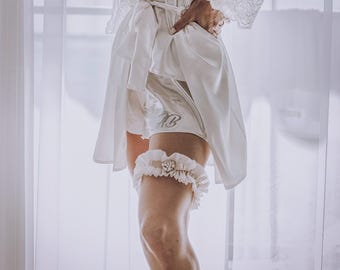 Könnte beinhalten: Elfenbeinfarbene Brautshorts aus Satin mit gestickten Initialen und einem passenden Strumpfband. Das Strumpfband hat ein gerüschtes Design und eine zentrale Verzierung. Ein weißer Satin-Bademantel mit Spitzenärmeln ist ebenfalls zu sehen.