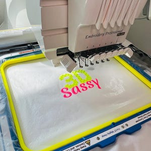 May include: A white embroidery machine with multiple thread spools and a needle head positioned over a yellow 11x13 inch hoop. The hoop secures a white fabric piece featuring the embroidered text 3D in lime green and Sassy in hot pink.