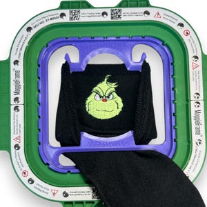 Könnte beinhalten: Ein Stickrahmen mit grünem Rahmen und lila Innenstütze hält einen schwarzen Stoff mit einem gestickten Grinch-Gesicht. Der Rahmen trägt die Aufschriften "MaggieFrame" und "3DSassy LLC". Die Innenabmessung beträgt 130 x 130 mm.