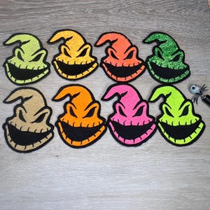 May include: A collection of eight colorful Oogie Boogie patches. Each patch features a smiling face with a stitched black mouth and eyes, and a pointed hat. The patches are in shades of green, orange, pink, and tan.