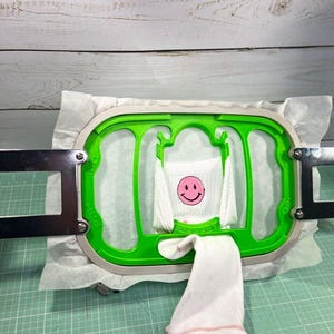 May include: A white sock featuring a pink smiley face is held in a green embroidery hoop. The hoop is attached to a metal frame, set on a green cutting mat. The background is a white wooden surface.