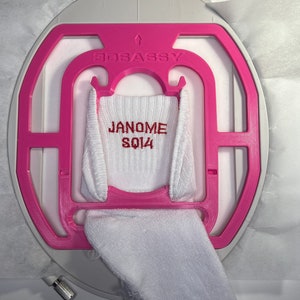 May include: A white sock is held in a pink embroidery hoop with the text "JANOME SQ14" embroidered in red. The hoop is labeled "3DSASSY" and is attached to a larger white and gray machine component.