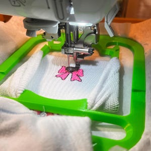 May include: A close-up of a white fabric with a pink bow being embroidered by a sewing machine. The machine has a green hoop holding the fabric in place. The needle is visible in the centre of the image.