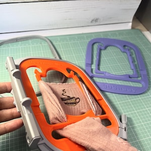 May include: An orange and gray embroidery hoop holds a pink fabric with a coffee cup design. A purple frame with the text "3DSassy" is in the background. The image is on a green cutting mat.