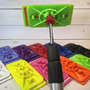 May include: A close-up of a green phone grip with a red button and a black selfie stick. The phone grip is attached to the selfie stick. There are multiple phone grips in different colors in the background.