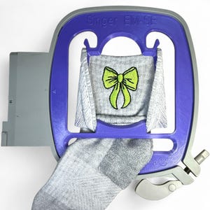 May include: A Singer EM-SE embroidery hoop holds a gray sock with a lime green bow design. The hoop is purple and gray, with the Singer logo at the top. The sock is partially visible, showing the embroidered bow.