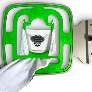 May include: A green embroidery hoop holds a white fabric with a black embroidered Highland cow design. The hoop has text that reads "Janome SQ 14b" and "ELNA 8.30". The fabric is partially visible, showing the embroidered design.