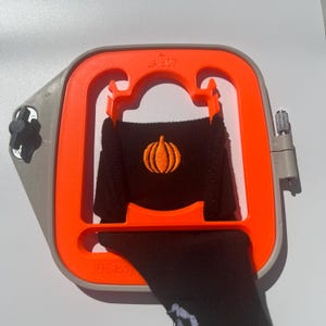 May include: An orange and gray embroidery hoop holds a black sock with an embroidered orange pumpkin design. The hoop has the text "JA807" and "3DSassy". The sock is partially visible, with the pumpkin design centered.