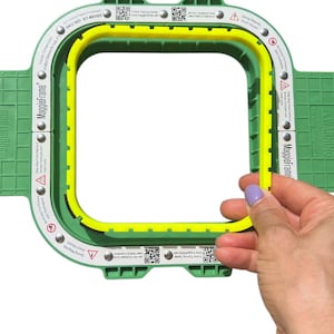 Op de afbeelding: Een groene en gele MaggieFrame borduurring. De ring heeft een vierkante vorm met afgeronde hoeken. De binnenafmetingen zijn 13 x 13 cm. De ring wordt vastgehouden door een hand met paarse nagellak.