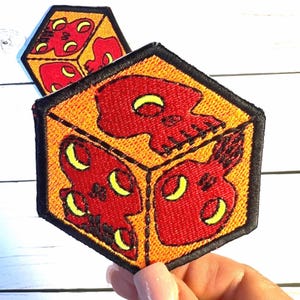 May include: Two embroidered patches in a hexagonal shape. Each patch features a red skull design on a yellow background, outlined in black. The skulls have yellow crescent-shaped eyes. One patch is in the background, and the other is in the foreground.