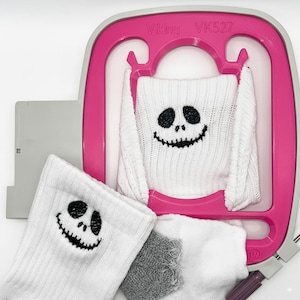 Puede incluir: Calcetines blancos con un diseño de cara negra de Jack Skellington se sostienen en un bastidor de bordado rosa. El bastidor tiene el texto "Viking VK527". También se ven piezas de tela blanca y gris adicionales.