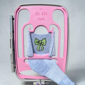 May include: A pink embroidery hoop with the text "SA 444 Adult" holds a light blue sock with a green bow design. The sock is partially embroidered and has a textured appearance. The hoop is open and ready for use.