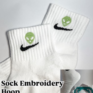 Pu&ograve; includere: Calzini bianchi alla caviglia con un swoosh Nike nero e un ricamo di alieno verde. I calzini sono in un cerchio da ricamo, con il testo "Sock Embroidery Hoop" sotto.