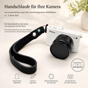 Könnte beinhalten: Eine schwarze Kamerahandschlaufe aus Büffelleder mit silbernem Metallniet und türkisfarbener Perle, befestigt an einer weißen Nikon 1 J3 Digitalkamera mit schwarzem Objektiv. Die Schlaufe ist 3,5 mm dick und besitzt einen Metall-Spaltring.