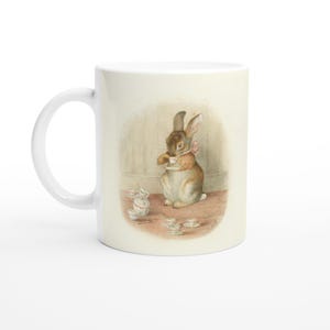May include: White ceramic mug with a watercolour illustration of a brown rabbit holding a teacup, with a pink bow. The design includes small teacups and a second rabbit. A charming, whimsical design.