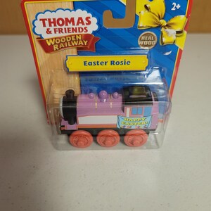 May include: A Thomas & Friends Wooden Railway Easter Rosie toy train in its original packaging. The train is lavender with a black roof, pink wheels, and a banner on the side that reads Happy Easter. The packaging features a yellow bow graphic.