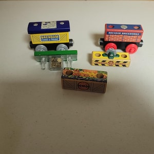 May include: Wooden toy train cars in various colors. One car is blue and yellow with "Brendam Cargo & Freight" text. Another is red and black with "Brendam Brickworks" text. A yellow car reads "Logging Station".